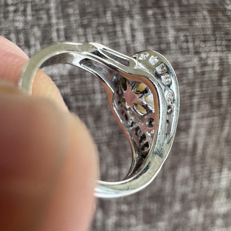May include: Silver ring with a heart-shaped design, featuring a central yellow stone and small clear stones along the edges. The ring is held by a person's finger, showcasing the intricate details of the setting.