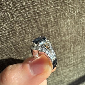 May include: A silver ring with a large, oval, dark blue gemstone as the centerpiece. The ring is encrusted with smaller, clear gemstones along the band and around the main stone, creating a sparkling effect. A small gold accent is visible.