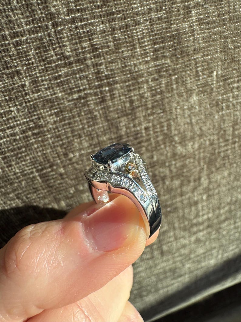 May include: A silver ring with a large, oval, dark blue gemstone as the centerpiece. The ring is encrusted with smaller, clear gemstones along the band and around the main stone, creating a sparkling effect. A small gold accent is visible.
