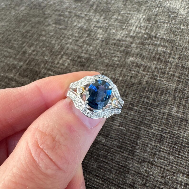 May include: A silver ring featuring a large, oval-cut, deep blue gemstone as the centerpiece. The ring is accented with smaller, clear gemstones and two small gold-colored stones. The setting has a decorative, open design.