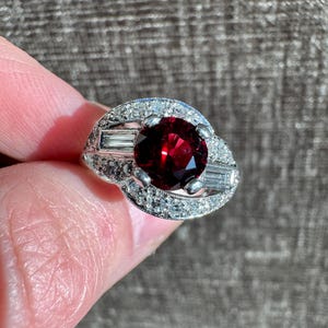 May include: A silver ring featuring a large, round, deep red gemstone as the focal point. The ring is surrounded by smaller, clear gemstones and baguette-cut stones, creating a halo effect. The ring is held up by a person's finger.