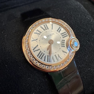 Puede incluir: Un reloj de pulsera de lujo con caja dorada y correa de cuero negro. La esfera del reloj presenta n&uacute;meros romanos y la marca "Cartier". El bisel est&aacute; incrustado con peque&ntilde;os diamantes. Una peque&ntilde;a gema azul est&aacute; engastada en el lateral.