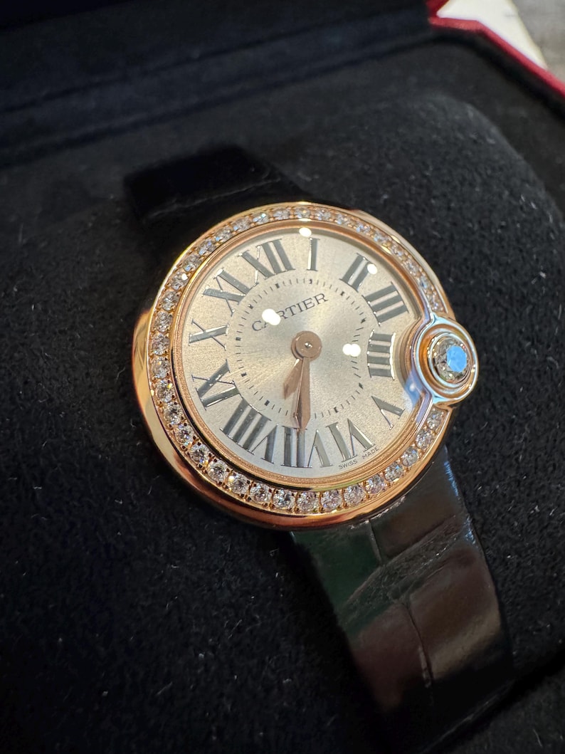 Puede incluir: Un reloj de pulsera de lujo con caja dorada y correa de cuero negro. La esfera del reloj presenta n&uacute;meros romanos y la marca "Cartier". El bisel est&aacute; incrustado con peque&ntilde;os diamantes. Una peque&ntilde;a gema azul est&aacute; engastada en el lateral.