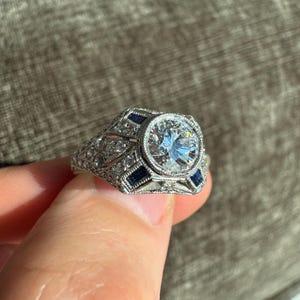 K&ouml;nnte beinhalten: Ein detaillierter Silberring mit einem gro&szlig;en, klaren, rund geschliffenen Mittelstein. Der Ring ist mit kleineren klaren Steinen und rechteckigen blauen Steinen besetzt, die in einem filigranen Design angeordnet sind.