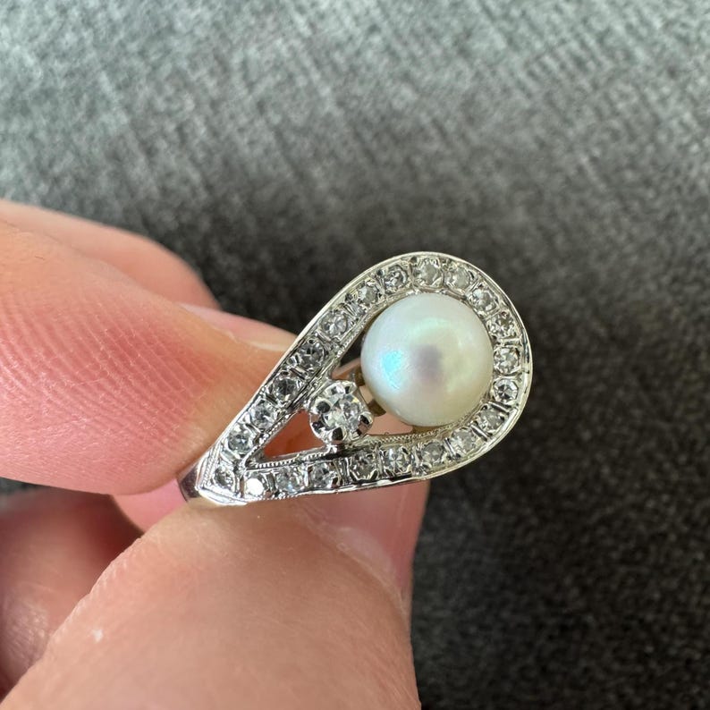 May include: A silver ring featuring a large, round pearl surrounded by small, clear gemstones. A smaller gemstone is set beside the pearl. The ring has a teardrop shape.