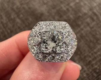 May include: A silver ring with a large, square-cut diamond center stone, surrounded by smaller diamonds in a halo and geometric design. The ring has an Art Deco style, with intricate detailing and a bright, reflective appearance.