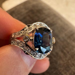 May include: A silver ring with a large, cushion-cut, deep blue gemstone as the centerpiece. The ring is encrusted with smaller, clear gemstones and gold accents. The ring is held up by a hand.
