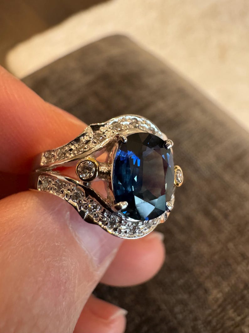 May include: A silver ring with a large, cushion-cut, deep blue gemstone as the centerpiece. The ring is encrusted with smaller, clear gemstones and gold accents. The ring is held up by a hand.