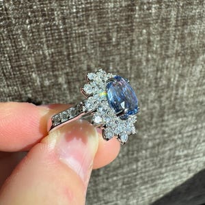 May include: A silver ring featuring a large, oval-cut, light blue gemstone surrounded by smaller, clear gemstones. The ring's band is also adorned with small, clear gemstones. The ring is held up against a neutral background.