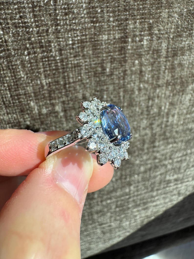 May include: A silver ring featuring a large, oval-cut, light blue gemstone surrounded by smaller, clear gemstones. The ring's band is also adorned with small, clear gemstones. The ring is held up against a neutral background.