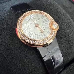 Puede incluir: Un reloj de lujo con una caja redonda dorada y una esfera blanca con n&uacute;meros romanos. El bisel est&aacute; incrustado con peque&ntilde;os diamantes. El reloj tiene una correa de cuero negro y la marca "CARTIER" es visible en la esfera.