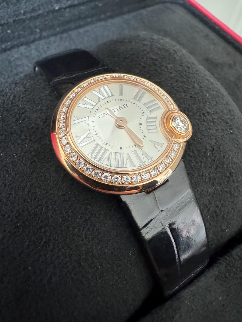 Puede incluir: Un reloj de lujo con una caja redonda dorada y una esfera blanca con n&uacute;meros romanos. El bisel est&aacute; incrustado con peque&ntilde;os diamantes. El reloj tiene una correa de cuero negro y la marca "CARTIER" es visible en la esfera.
