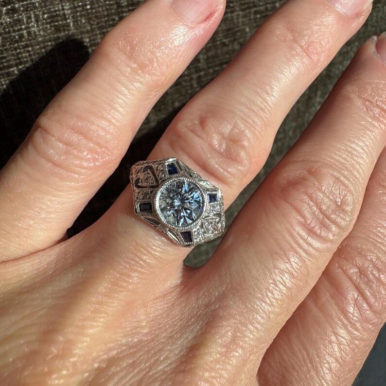 K&ouml;nnte beinhalten: Ein Ring im Vintage-Stil mit einem gro&szlig;en, runden, hellblauen Edelstein als Mittelpunkt. Der Ring weist aufwendige Filigranarbeiten auf und ist mit kleineren, dunkelblauen Edelsteinen verziert. Die Fassung ist silberfarben und bildet einen auff&auml;lligen Kontrast zu den Steinen.