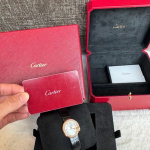 Puede incluir: Un reloj Cartier con correa de cuero negro y bisel dorado con esfera con diamantes. El reloj se exhibe sobre un coj&iacute;n de terciopelo negro dentro de una caja negra. Tambi&eacute;n hay una caja y una tarjeta rojas con el logotipo de Cartier.