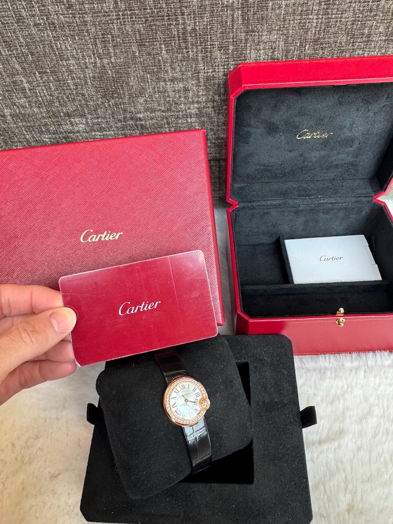 Puede incluir: Un reloj Cartier con correa de cuero negro y bisel dorado con esfera con diamantes. El reloj se exhibe sobre un coj&iacute;n de terciopelo negro dentro de una caja negra. Tambi&eacute;n hay una caja y una tarjeta rojas con el logotipo de Cartier.