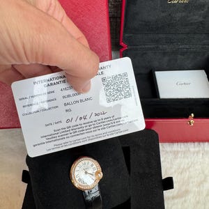 Puede incluir: Un reloj Cartier Ballon Blanc en oro rosa con correa de cuero negro. El reloj tiene una esfera blanca con n&uacute;meros romanos y un bisel con diamantes. Se muestra en una caja de terciopelo negro, con una caja roja de Cartier en el fondo.