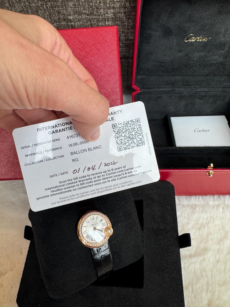 Puede incluir: Un reloj Cartier Ballon Blanc en oro rosa con correa de cuero negro. El reloj tiene una esfera blanca con n&uacute;meros romanos y un bisel con diamantes. Se muestra en una caja de terciopelo negro, con una caja roja de Cartier en el fondo.