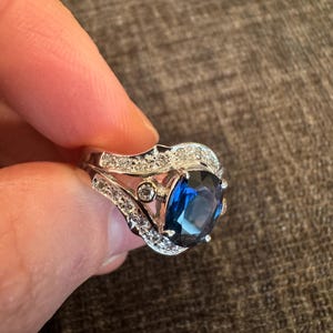 May include: A silver ring with a large, oval, deep blue gemstone as the centerpiece. The ring is adorned with smaller, clear gemstones along the band and around the main stone, creating a sparkling effect. The ring is held by a person.