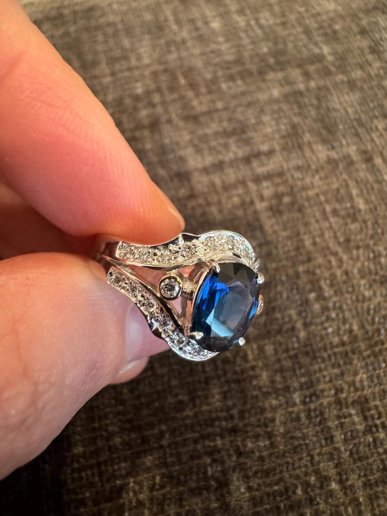 May include: A silver ring with a large, oval, deep blue gemstone as the centerpiece. The ring is adorned with smaller, clear gemstones along the band and around the main stone, creating a sparkling effect. The ring is held by a person.