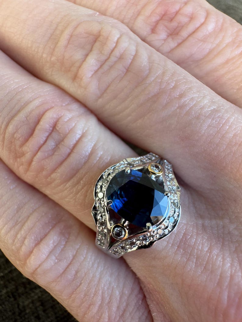 May include: A silver ring featuring a large, oval-cut, deep blue gemstone as the centerpiece. The ring is adorned with smaller, clear gemstones and gold accents, creating a luxurious and elegant design. The ring is a statement piece.