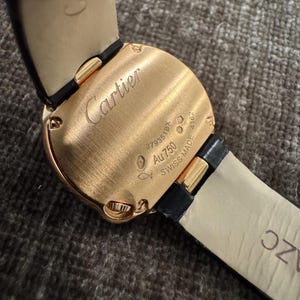 Puede incluir: Primer plano de una caja de reloj Cartier en oro rosa con una correa de cuero negro. La caja del reloj tiene el logotipo de Cartier grabado, junto con el texto "Au750" y "SWISS MADE".