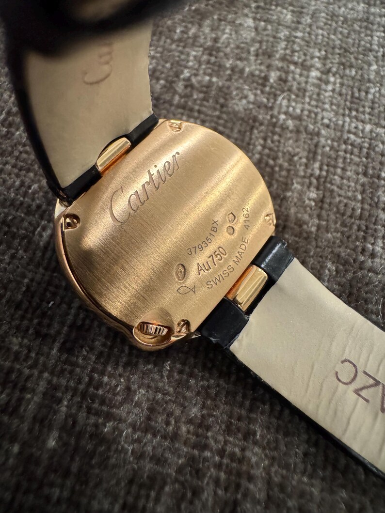 Puede incluir: Primer plano de una caja de reloj Cartier en oro rosa con una correa de cuero negro. La caja del reloj tiene el logotipo de Cartier grabado, junto con el texto "Au750" y "SWISS MADE".