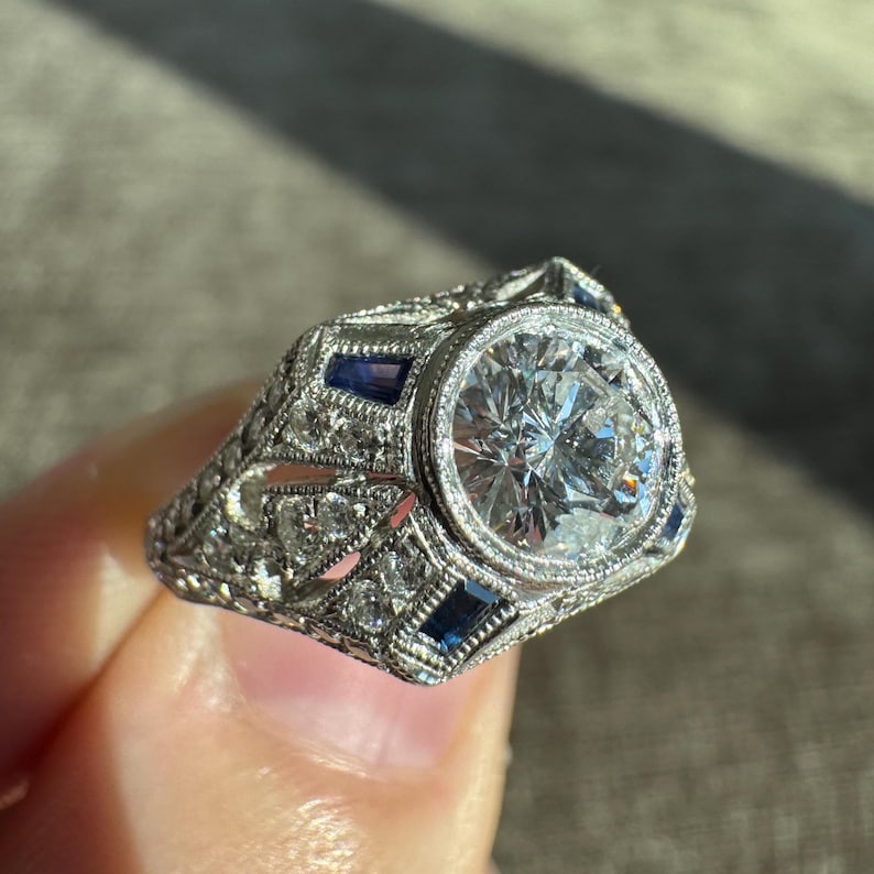 K&ouml;nnte beinhalten: Ein silberner Ring mit einem gro&szlig;en, runden, klaren Edelstein in der Mitte, umgeben von kleineren klaren Steinen und rechteckigen blauen Edelsteinen. Der Ring hat ein verziertes Filigran-Design.