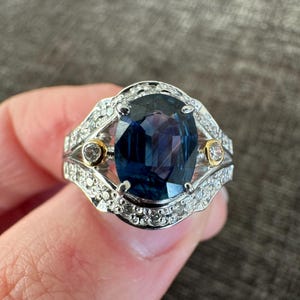 May include: A silver ring with a large, oval-cut, deep blue gemstone as the centerpiece. The ring is accented with smaller, clear gemstones and two gold-colored accents. The setting is intricate and detailed, showcasing a classic jewelry design.