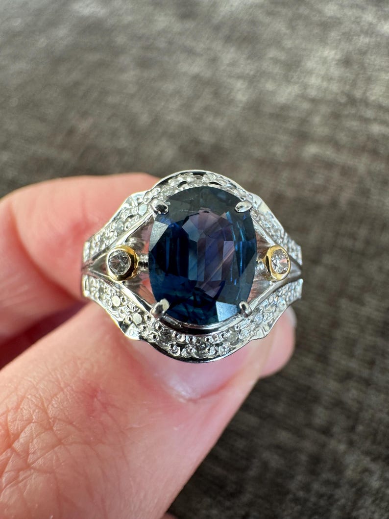 May include: A silver ring with a large, oval-cut, deep blue gemstone as the centerpiece. The ring is accented with smaller, clear gemstones and two gold-colored accents. The setting is intricate and detailed, showcasing a classic jewelry design.