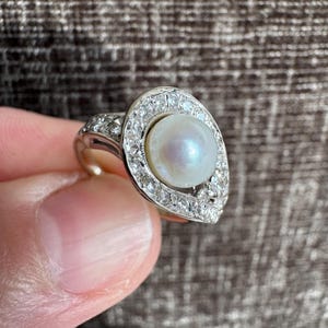 May include: A vintage-style ring featuring a large, lustrous pearl at the center, surrounded by a halo of small, sparkling diamonds. The ring has an ornate, detailed setting, likely made of white gold or platinum, and is a classic piece of jewelry.