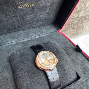 Puede incluir: Un reloj Cartier de oro rosa con una correa de cuero negro descansa en una caja de terciopelo negro. La esfera del reloj tiene n&uacute;meros romanos y est&aacute; rodeada de peque&ntilde;os diamantes. El logotipo de Cartier es visible en la caja.
