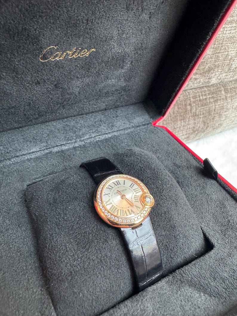 Puede incluir: Un reloj Cartier de oro rosa con una correa de cuero negro descansa en una caja de terciopelo negro. La esfera del reloj tiene n&uacute;meros romanos y est&aacute; rodeada de peque&ntilde;os diamantes. El logotipo de Cartier es visible en la caja.