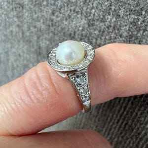 May include: A silver ring featuring a large, round, white pearl center stone, surrounded by a halo of small, clear gemstones. The ring's band is also adorned with smaller gemstones.