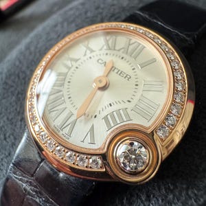 Puede incluir: Reloj de lujo con caja de oro rosa y esfera blanca con n&uacute;meros romanos. El reloj tiene un bisel con diamantes y un diamante grande a las 6. Se presenta sobre una correa de cuero negro.