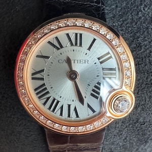 Puede incluir: Un reloj de pulsera de lujo con esfera plateada, n&uacute;meros romanos negros y bisel de oro rosa con diamantes incrustados. El reloj presenta la marca "CARTIER" y un peque&ntilde;o acento de diamante. Tiene una correa de cuero negro.
