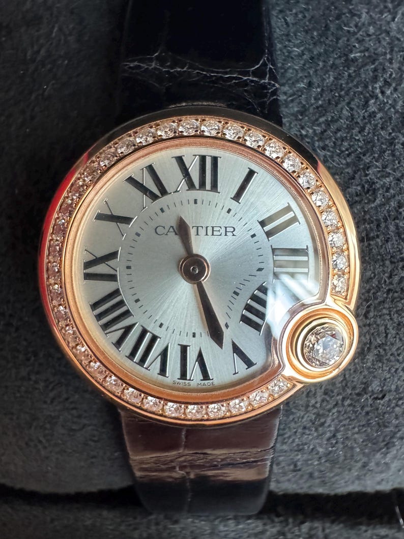 Puede incluir: Un reloj de pulsera de lujo con esfera plateada, n&uacute;meros romanos negros y bisel de oro rosa con diamantes incrustados. El reloj presenta la marca "CARTIER" y un peque&ntilde;o acento de diamante. Tiene una correa de cuero negro.