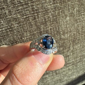 May include: A silver ring with a large, oval-cut, dark blue gemstone as the centerpiece. The ring is adorned with smaller, clear gemstones and intricate detailing along the band, creating a luxurious and elegant appearance. The ring is held up by a person's fingers.
