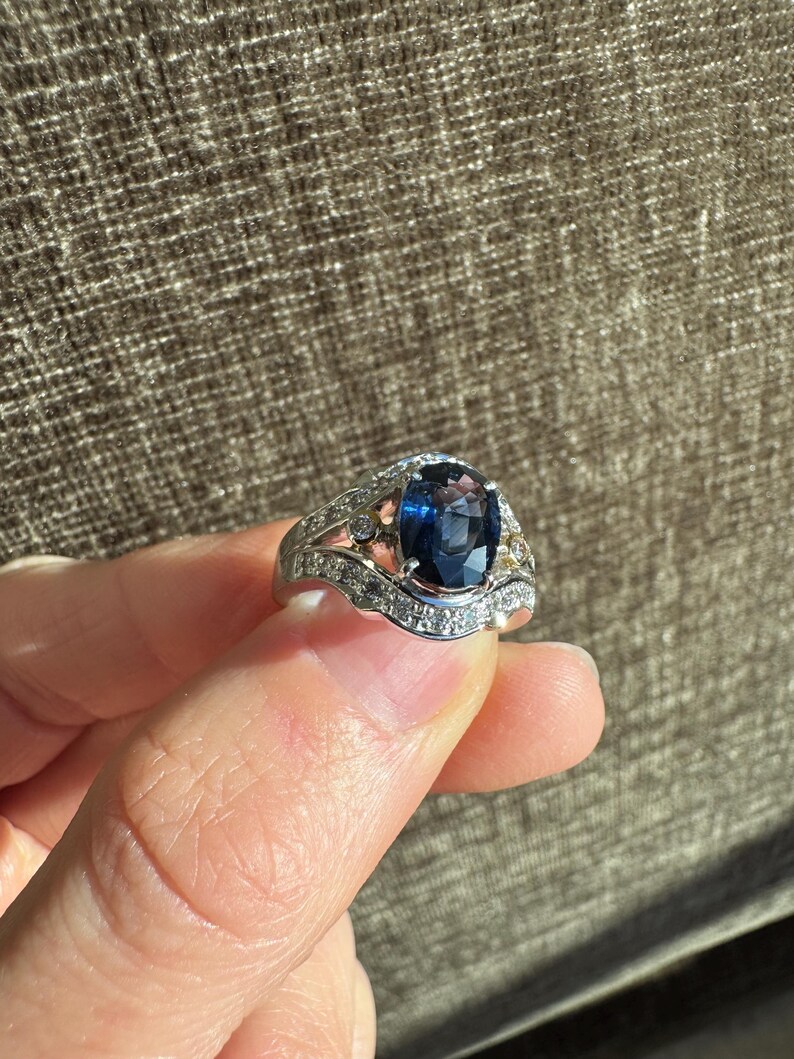 May include: A silver ring with a large, oval-cut, dark blue gemstone as the centerpiece. The ring is adorned with smaller, clear gemstones and intricate detailing along the band, creating a luxurious and elegant appearance. The ring is held up by a person's fingers.