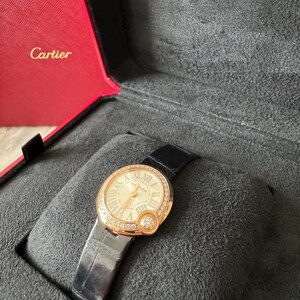 Puede incluir: Un reloj de pulsera dorado con una correa de cuero negro, presentado en un estuche rojo y gris. El reloj tiene una esfera redonda con n&uacute;meros romanos y un bisel con diamantes. La marca "Cartier" es visible.