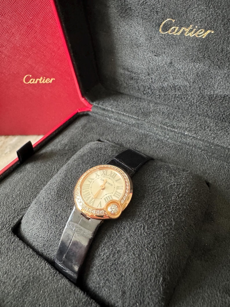 Puede incluir: Un reloj de pulsera dorado con una correa de cuero negro, presentado en un estuche rojo y gris. El reloj tiene una esfera redonda con n&uacute;meros romanos y un bisel con diamantes. La marca "Cartier" es visible.