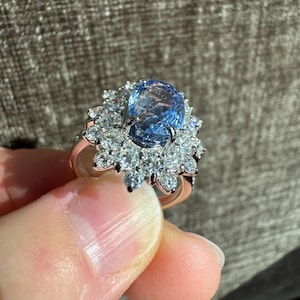 May include: A rose gold ring featuring a large, oval-cut, light blue gemstone surrounded by smaller, clear gemstones. The ring's design is a cluster of sparkling stones, creating a halo effect around the central gem. The ring is held up by a person's fingers.