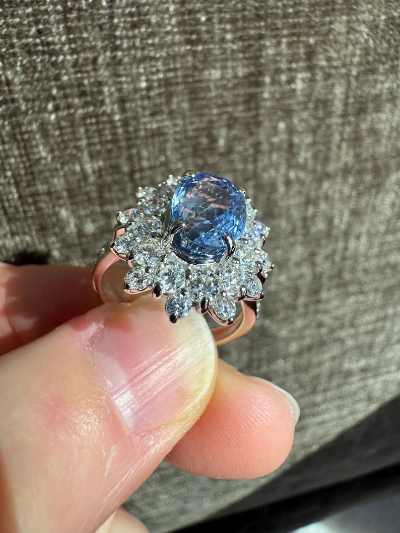 May include: A rose gold ring featuring a large, oval-cut, light blue gemstone surrounded by smaller, clear gemstones. The ring's design is a cluster of sparkling stones, creating a halo effect around the central gem. The ring is held up by a person's fingers.