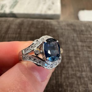 May include: A silver ring with a large, oval, dark blue gemstone as the centerpiece. The ring is adorned with smaller, clear gemstones and gold accents, creating a luxurious and elegant appearance.