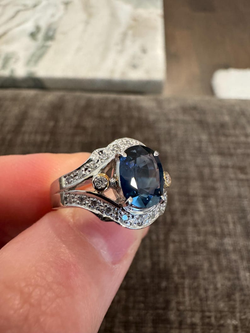 May include: A silver ring with a large, oval, dark blue gemstone as the centerpiece. The ring is adorned with smaller, clear gemstones and gold accents, creating a luxurious and elegant appearance.