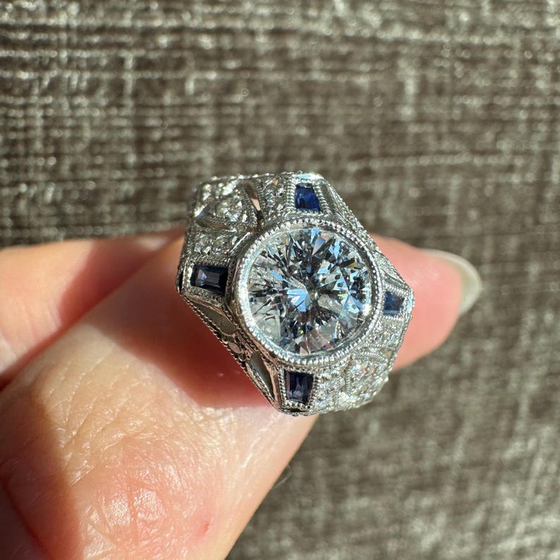 K&ouml;nnte beinhalten: Ein detaillierter, silberfarbener Ring mit einem gro&szlig;en, runden, klaren Edelstein im Zentrum. Der Ring ist mit kleineren, quadratischen, dunkelblauen Edelsteinen und aufwendigen Filigranarbeiten verziert. Ein klassisches, elegantes Design.