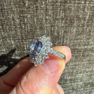 May include: A silver ring featuring a large, oval-cut blue gemstone surrounded by a halo of smaller, clear gemstones. The ring's band is also adorned with small, clear gemstones. The ring is held up against a brown background.