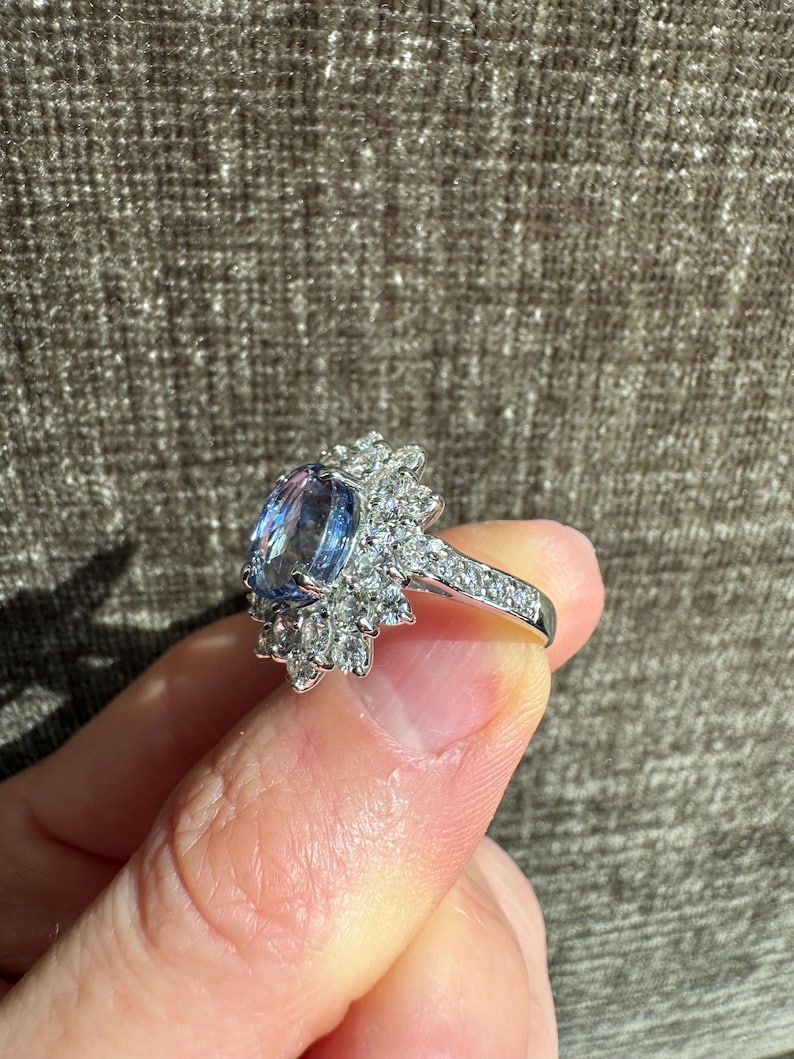 May include: A silver ring featuring a large, oval-cut blue gemstone surrounded by a halo of smaller, clear gemstones. The ring's band is also adorned with small, clear gemstones. The ring is held up against a brown background.