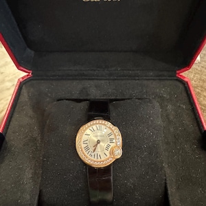 Puede incluir: Un reloj Cartier dorado con esfera redonda, n&uacute;meros romanos y bisel con diamantes. El reloj tiene una correa de cuero negro y se muestra en una caja roja y negra.