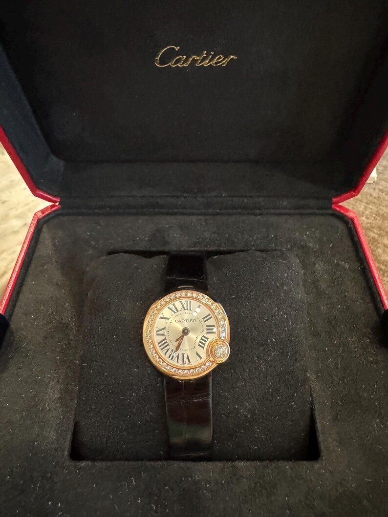 Puede incluir: Un reloj Cartier dorado con esfera redonda, n&uacute;meros romanos y bisel con diamantes. El reloj tiene una correa de cuero negro y se muestra en una caja roja y negra.