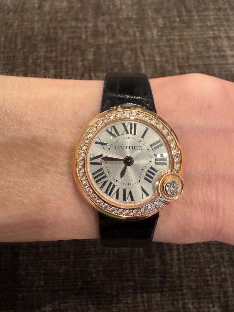 Puede incluir: Un reloj de pulsera dorado con esfera plateada y n&uacute;meros romanos negros. El reloj presenta un bisel con incrustaciones de diamantes y una correa de cuero negro. La palabra "CARTIER" est&aacute; impresa en la esfera. Hecho en Suiza.